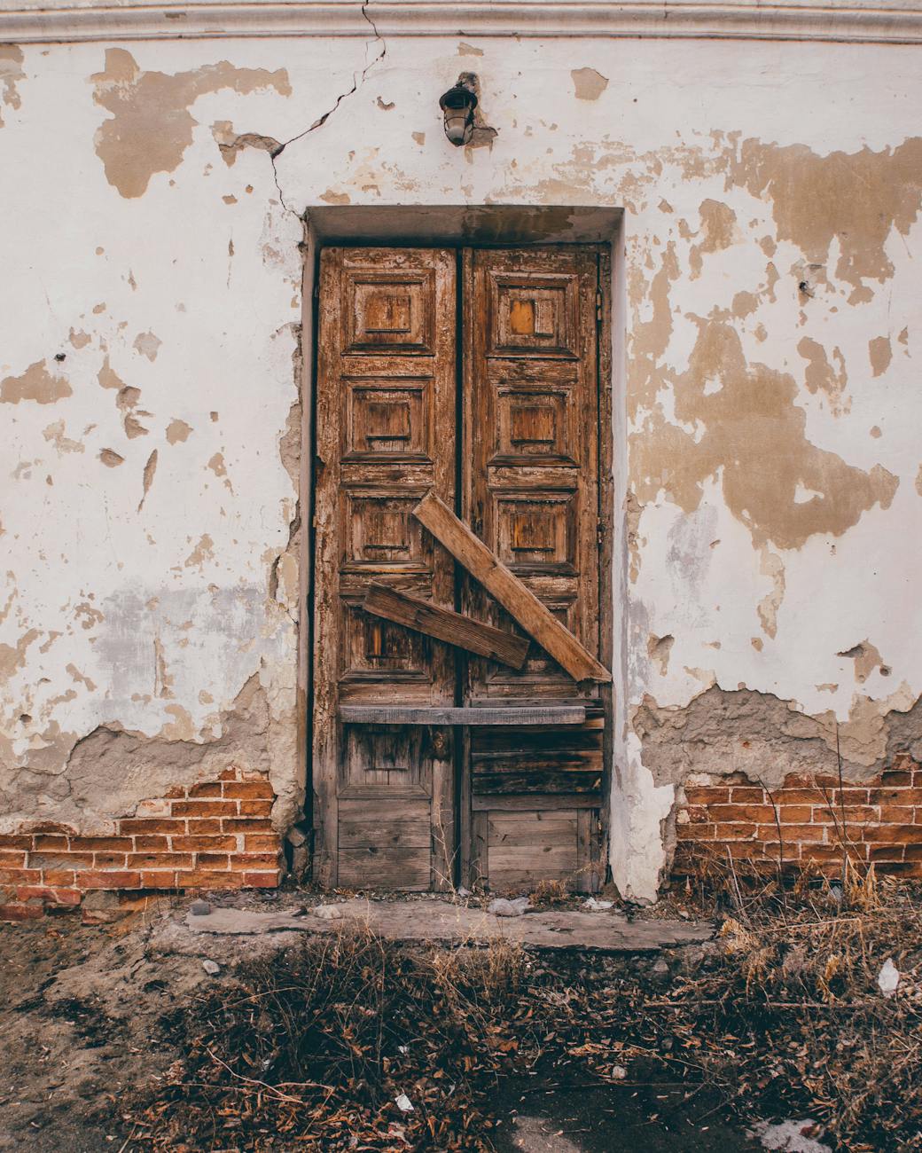 A photograph of a cracked wall and a broken door. The ground appears to be wasteland with dried grass and dried fallen branches. 