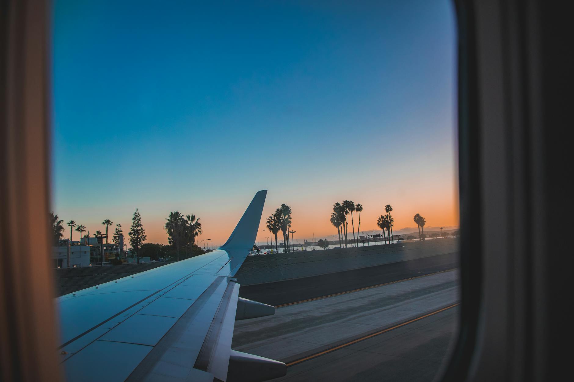A photograph of the wings of an airplane from inside the window of the airplane. The plane is on the runway taking off. There are some trees on the sides of the runway.