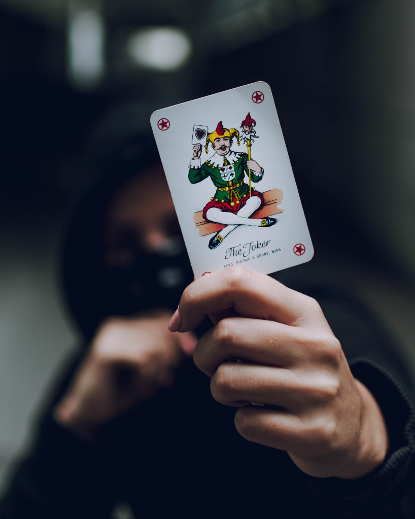 A photograph of a hoodie wearing man holding a Joker card in his hand. 